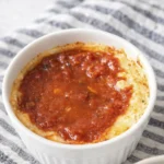 White ramekin filled with baked melted cheese topped with rich tomato sauce on a striped cloth background