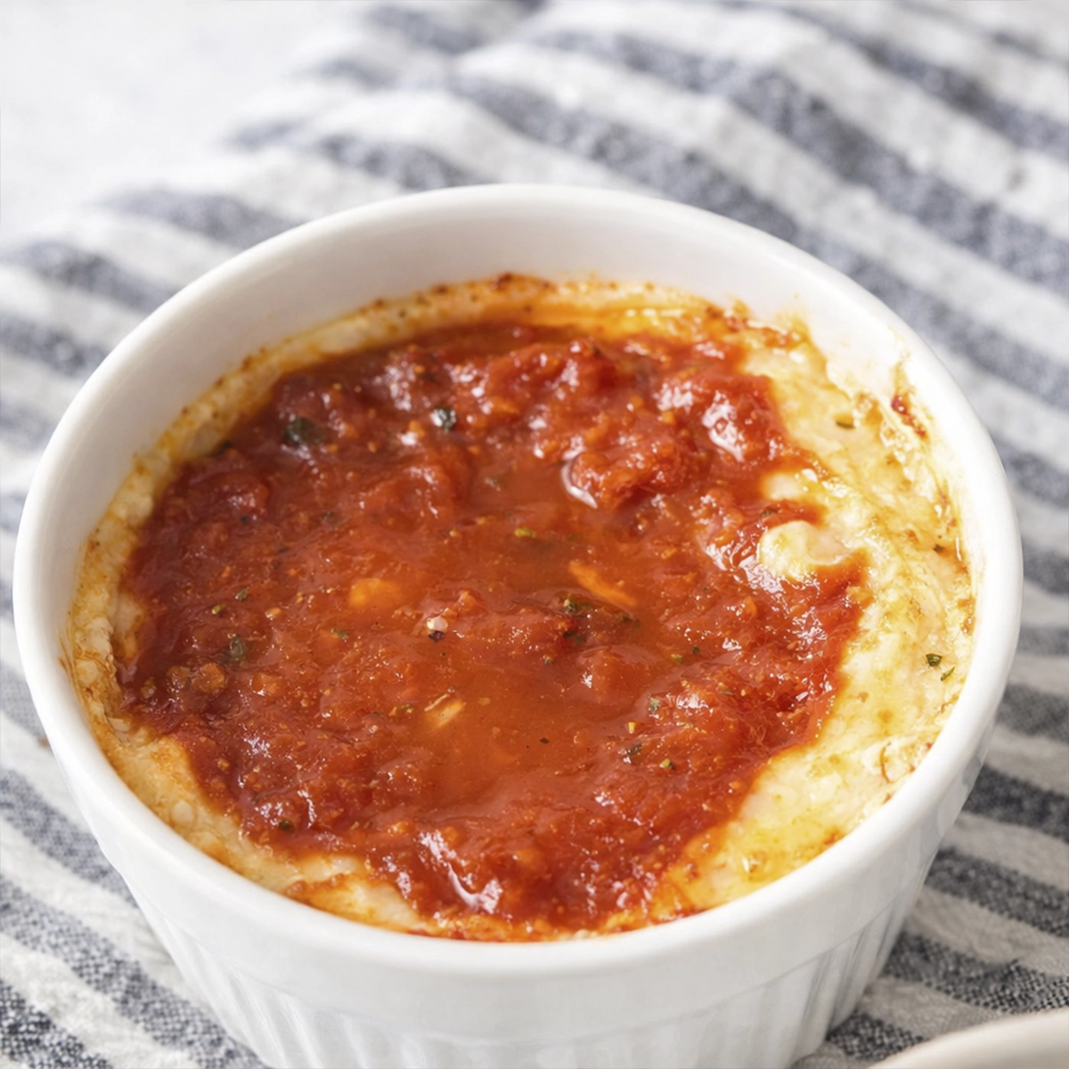 White ramekin filled with baked melted cheese topped with rich tomato sauce on a striped cloth background