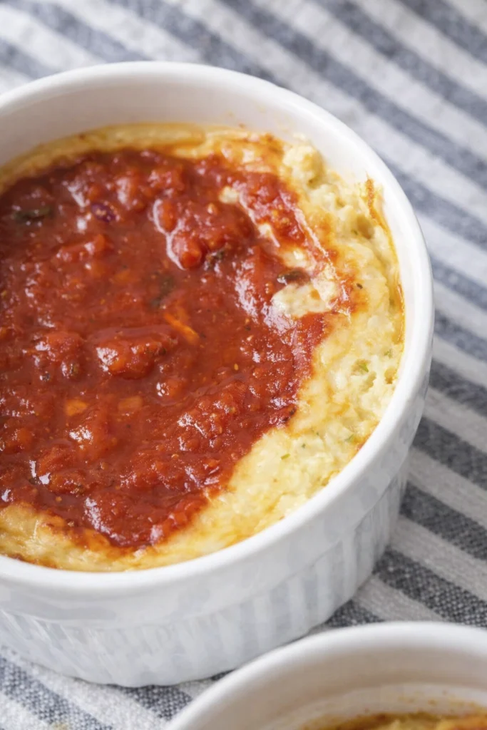 Close-up of creamy baked ricotta or custard in a white ramekin topped with chunky tomato sauce.