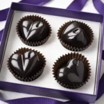 Four glossy heart-shaped dark chocolate truffles arranged in a white gift box with a purple ribbon, photographed from above on a clean background