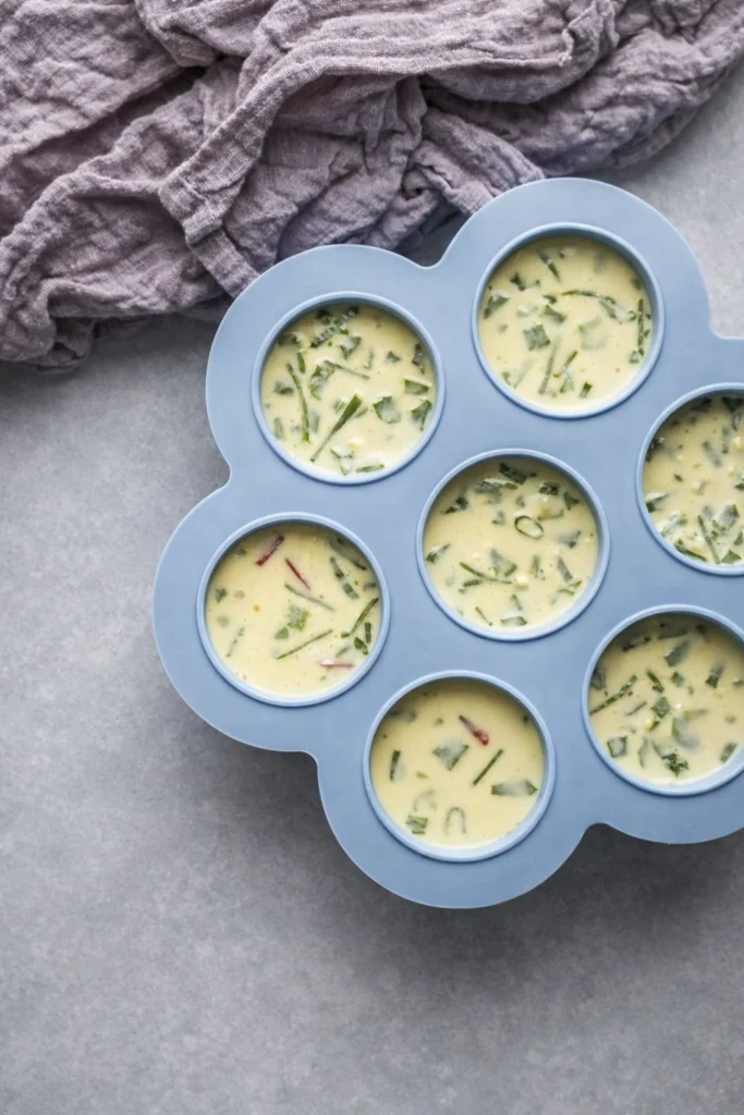 Raw egg mixture with herbs and vegetables in a blue silicone mold, ready for baking, top-down view