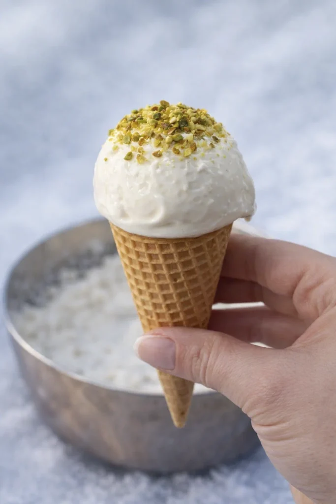 Hand holding a vanilla ice cream cone topped with crushed pistachios with a soft blurred background