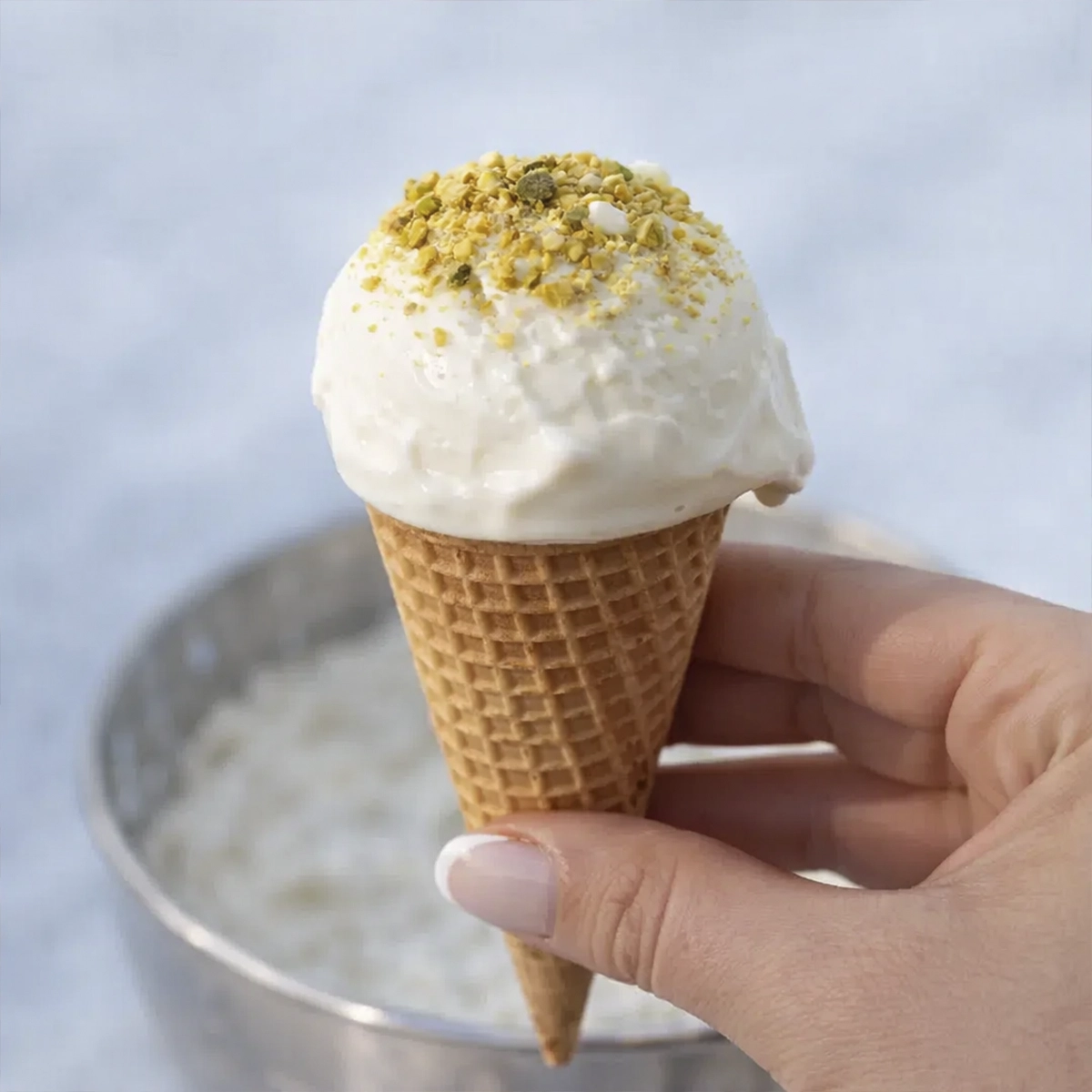 Hand holding a vanilla ice cream cone topped with crushed pistachios against a soft background