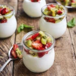 Creamy vanilla yogurt in glass jars topped with fresh strawberries and kiwi on a rustic wooden table.