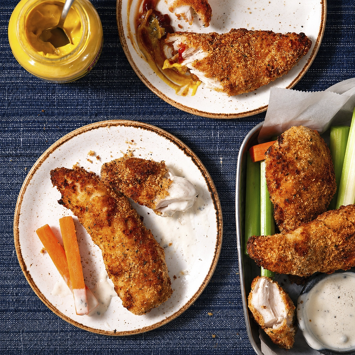 Breaded chicken tender being coated in seasoned crumbs beside egg mixture on a wooden table
