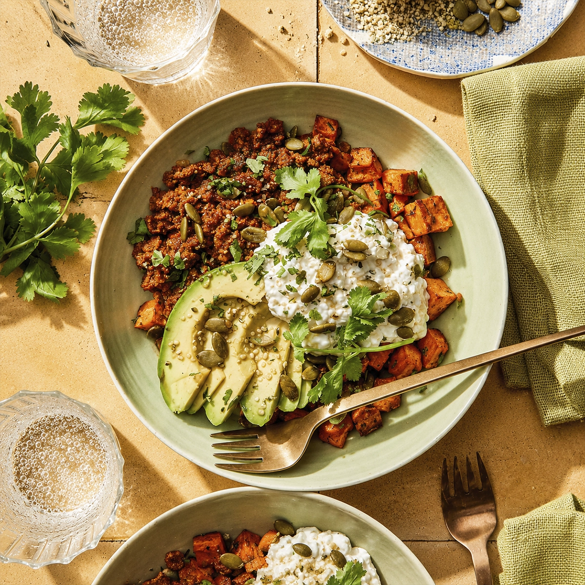 Healthy vegan nourish bowl with roasted sweet potatoes, avocado slices, creamy yogurt sauce, spicy plant-based crumble, pumpkin seeds, and fresh cilantro in warm natural lighting.