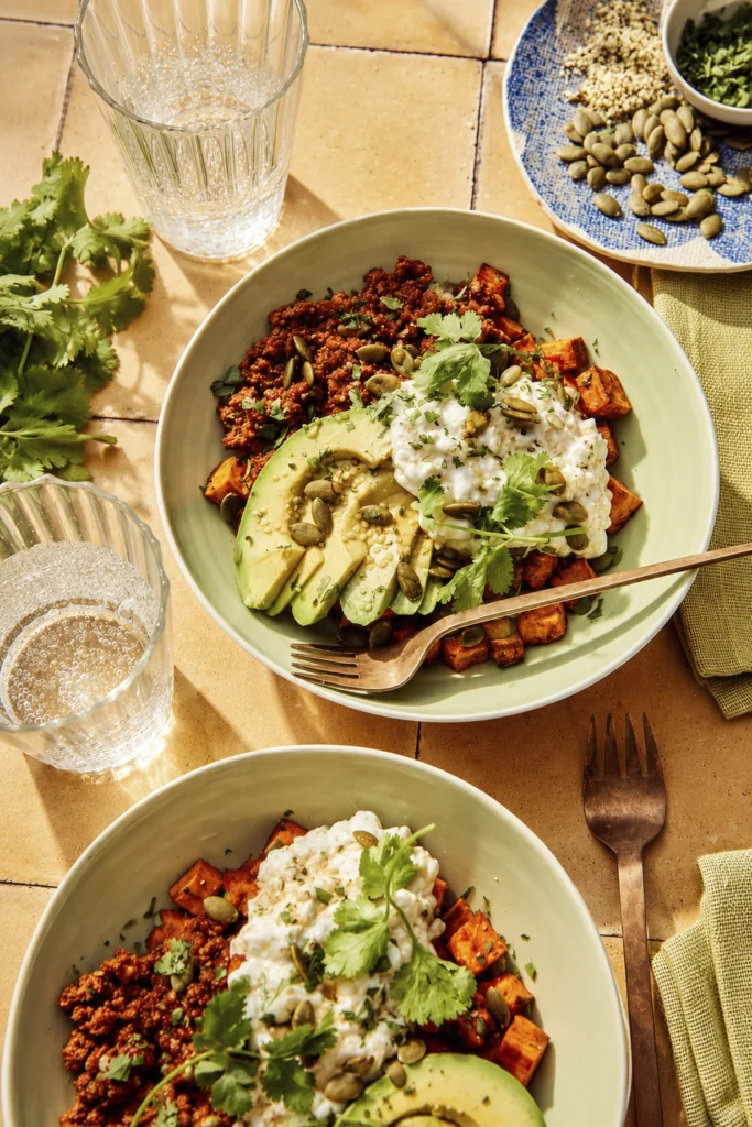 Overhead view of healthy vegan nourish bowls with roasted sweet potatoes, avocado, yogurt topping, pumpkin seeds, and cilantro on a warm rustic table setting.