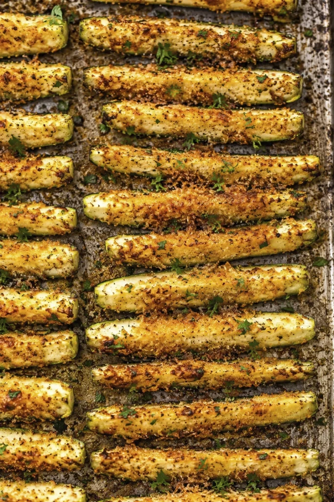 Crispy baked zucchini fries coated with Parmesan cheese and roasted until golden brown on a baking sheet with fresh parsley garnish.
