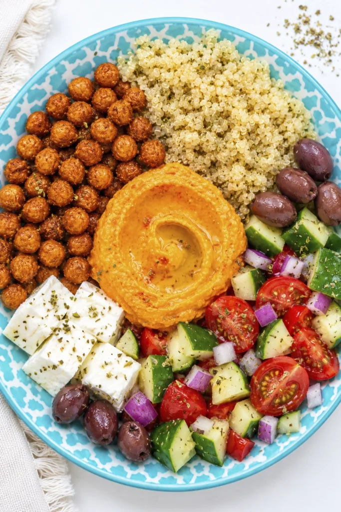 Top-down view of a healthy Mediterranean quinoa bowl with hummus, roasted chickpeas, feta cheese, fresh cucumber tomato salad, kalamata olives, and quinoa served on a turquoise patterned plate.