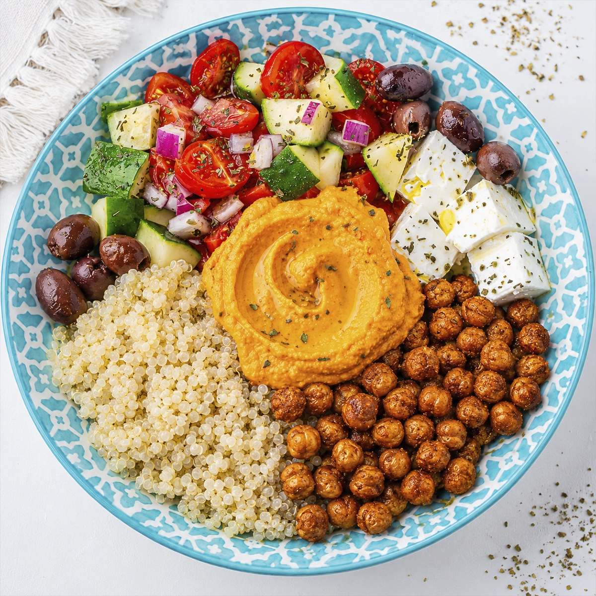 Mediterranean quinoa bowl with roasted chickpeas, hummus, feta cheese, Kalamata olives, and fresh Greek salad served in a turquoise patterned bowl.