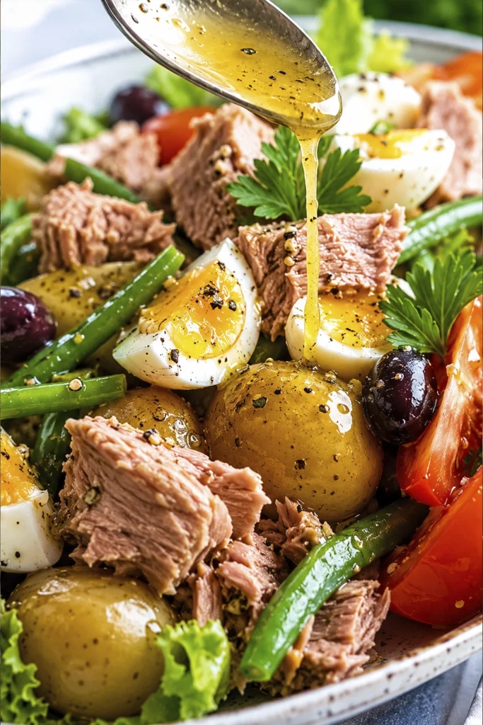 Close-up of a fresh Niçoise salad with tuna, baby potatoes, green beans, soft-boiled eggs, tomatoes, black olives, and lemon vinaigrette being poured over the salad.