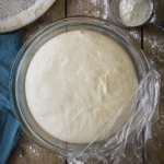 Freshly risen bagel dough resting in a glass mixing bowl covered with plastic wrap on a floured wooden table.