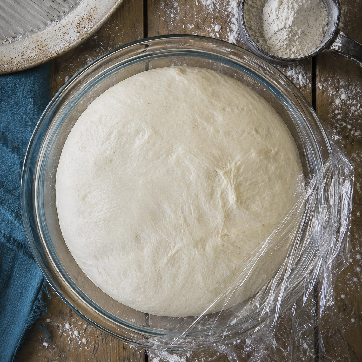 Freshly risen bagel dough resting in a glass mixing bowl covered with plastic wrap on a floured wooden table.