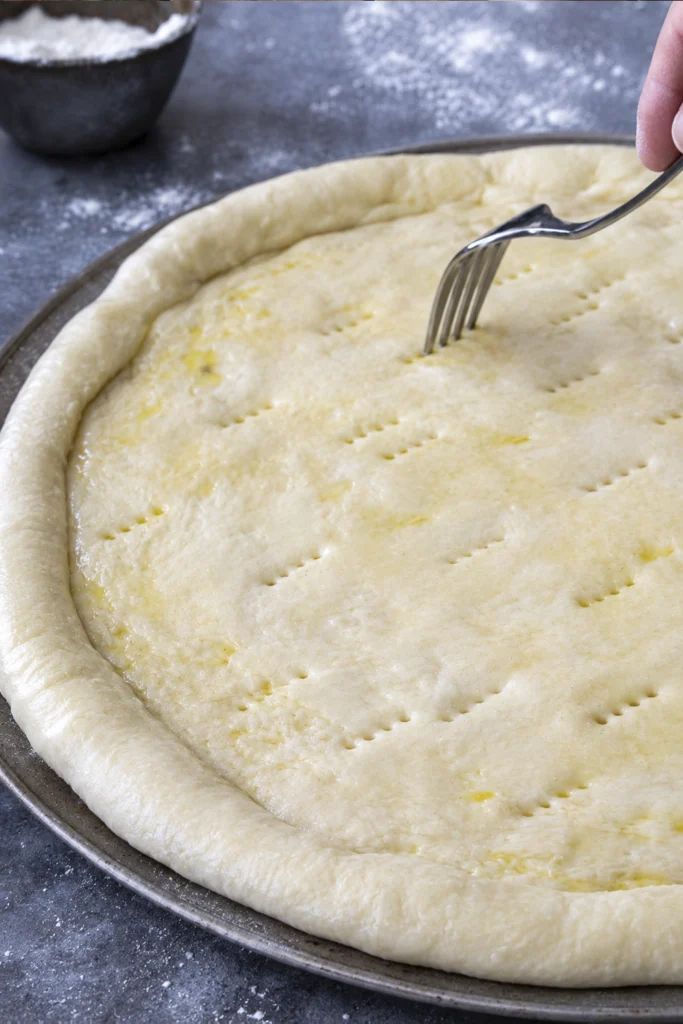 Fork docking homemade pizza dough in a round pizza pan before baking to prevent air bubbles.