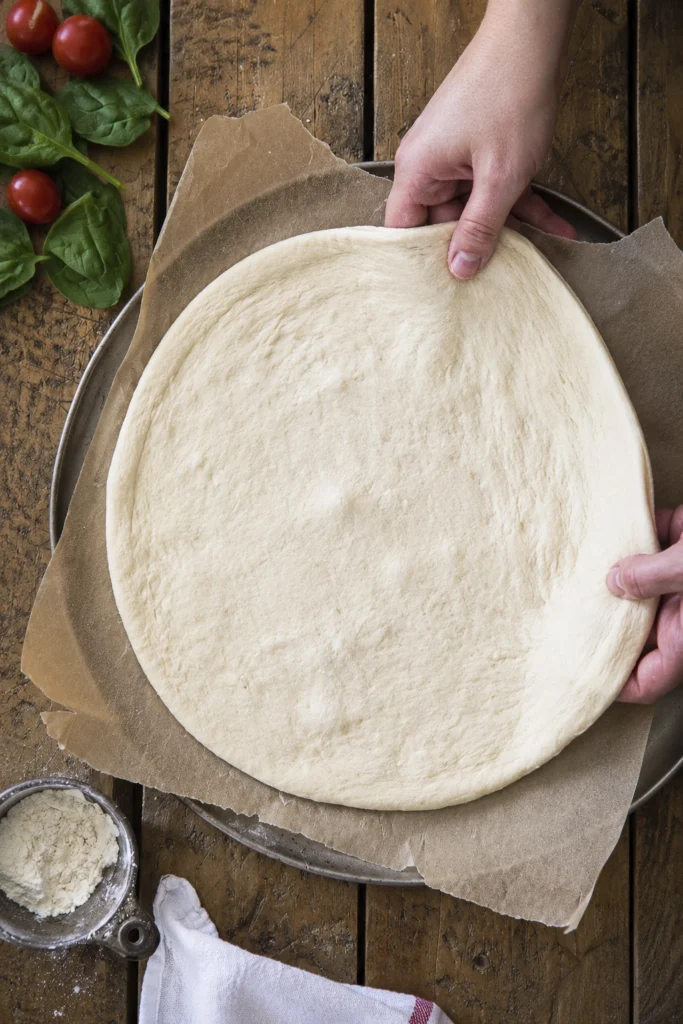 Hands stretching homemade pizza dough into a round crust on parchment paper over a pizza pan with fresh ingredients nearby.