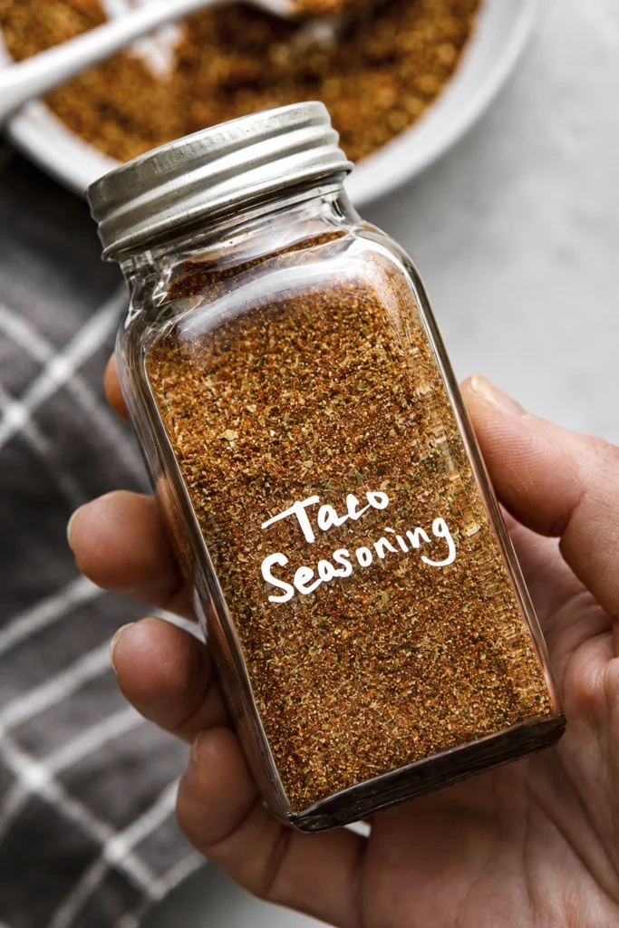 Hand holding a glass jar filled with homemade taco seasoning made from chili powder, paprika, cumin, garlic powder, and herbs.