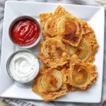 Golden crispy cheddar cheese crisps topped with caramelized onion slices, served on a white plate with ketchup and ranch dipping sauces.