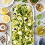 Overhead view of a fresh zucchini ribbon salad with basil, toasted pine nuts, lemon vinaigrette, and Parmesan on a white serving platter.