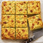 Golden baked zucchini breakfast casserole cut into squares with cheddar cheese, bacon, and fluffy eggs on a baking tray.