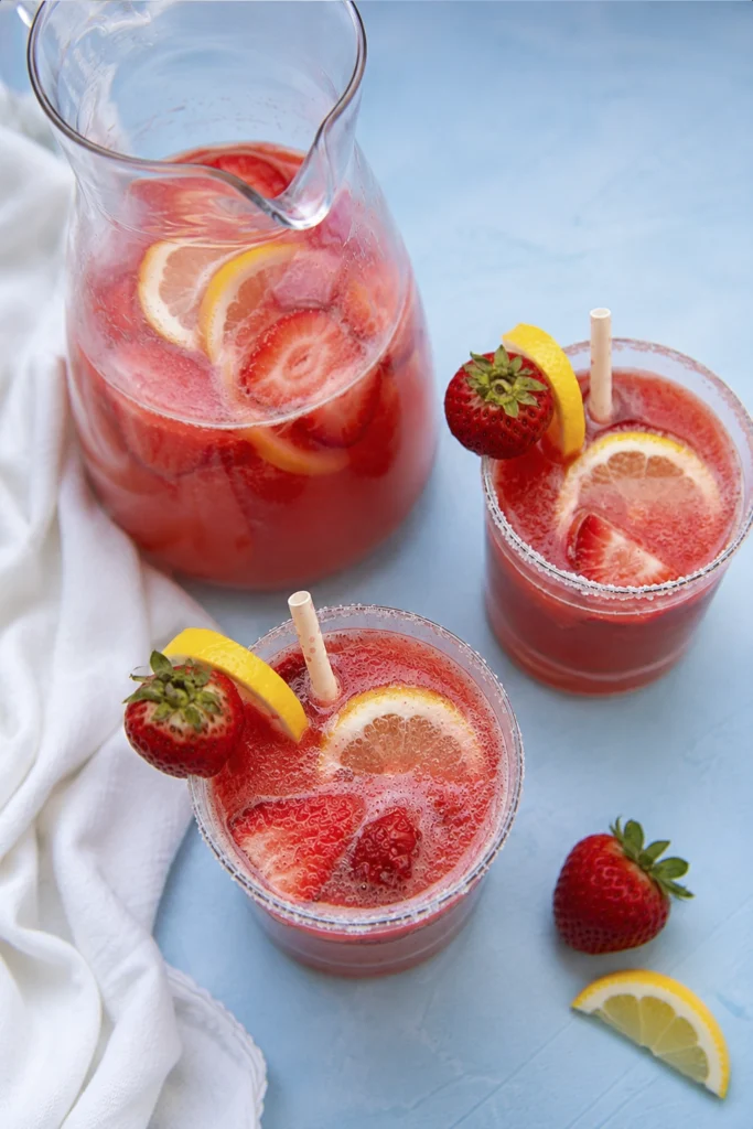Fresh summer drink in a pitcher with two garnished glasses, strawberries, lemon slices, and a soft blue background.