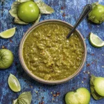 Homemade roasted tomatillo salsa verde in a rustic bowl with fresh tomatillos, lime wedges, and a spoon on a blue wooden table.