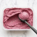 Creamy homemade mixed berry frozen yogurt with a fresh scoop and dark berry flecks in a freezer container.