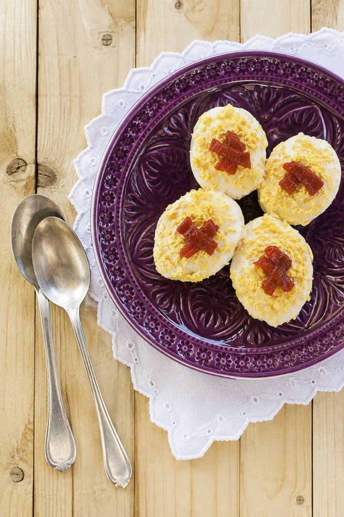 Stuffed hard-boiled eggs topped with grated yolk and roasted red pepper on a decorative purple plate with vintage silver spoons.