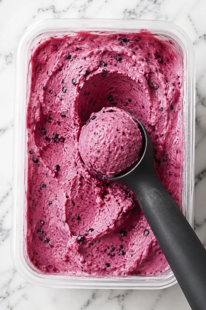 Homemade mixed berry frozen yogurt with a creamy berry scoop in a container on a white marble surface.