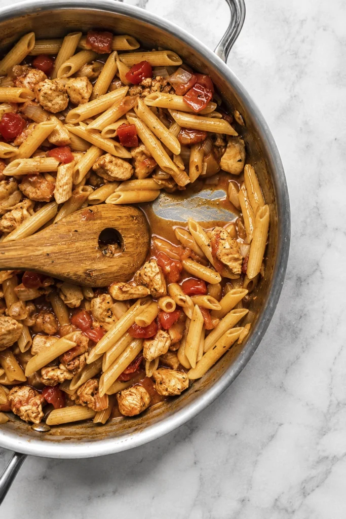 Cajun chicken penne pasta with tomatoes and onions cooking in a stainless-steel skillet.